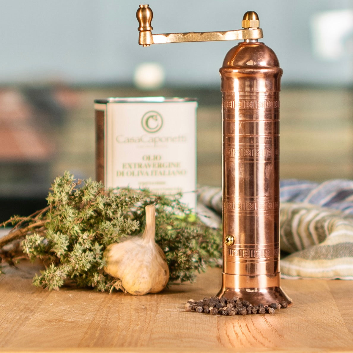 Copper pepper grinder on table with garlic and herbs and olive oil in the background